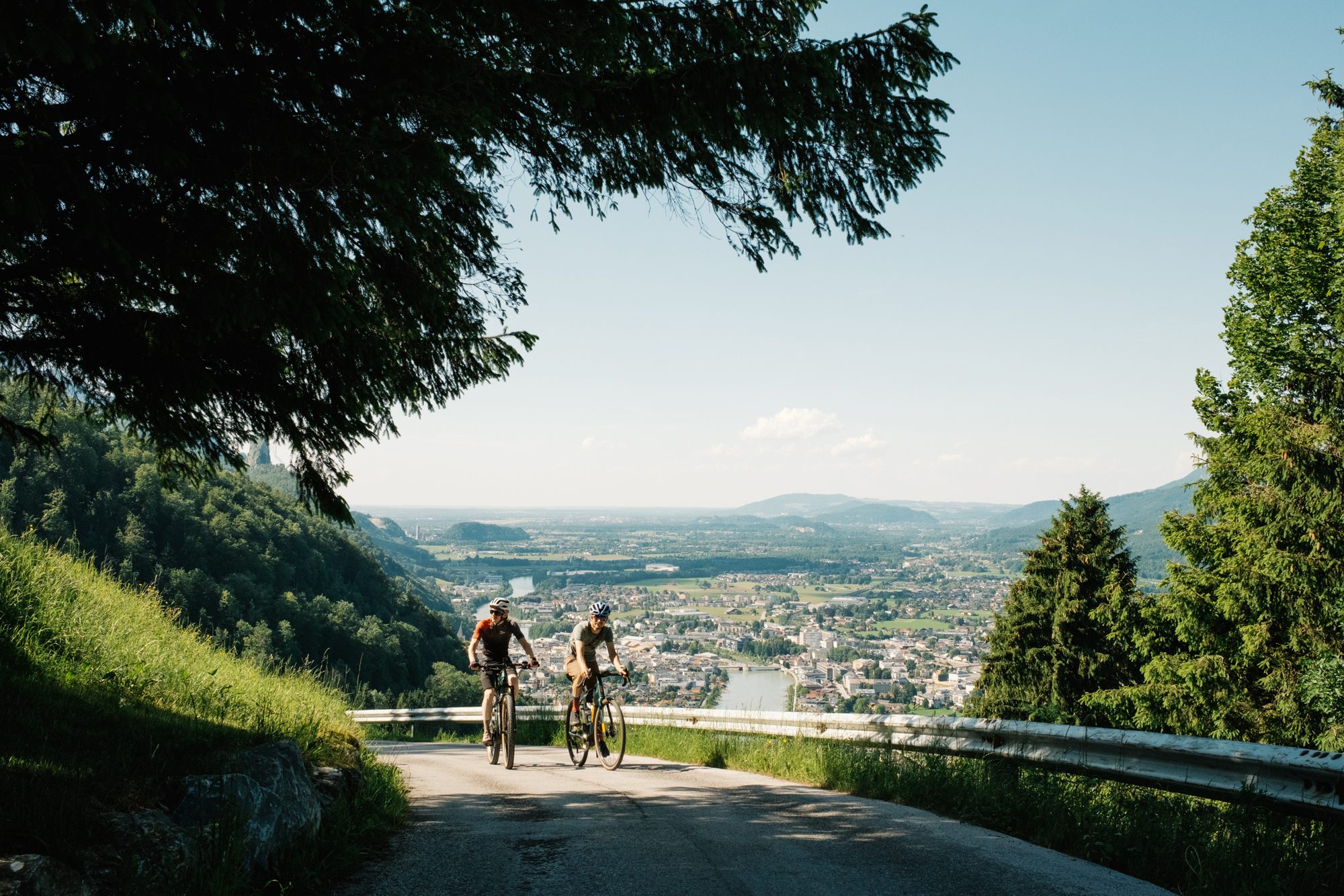 Zwei Fahrradfahrer fahren den Dürrnberg in Hallein hinauf. Im Hintergrund sieht man die Salzach, die Stadt Hallein und Teile Salzburgs